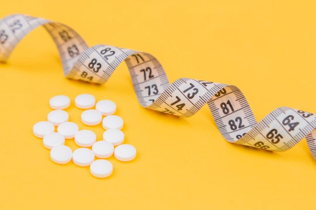 White tablets next to a coiled measuring tape on a yellow background, symbolizing medications and their impact on surgical outcomes and body regulation.